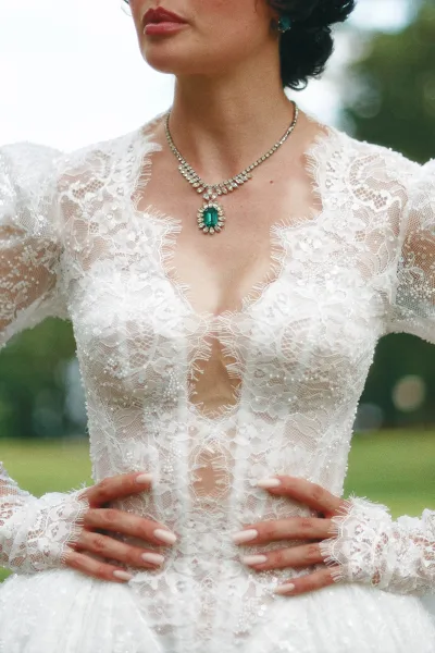 Lace wedding dress, a long sleeve bridal gown highlighted by an emerald pendant necklace, framed by garden trees over a grass lawn.