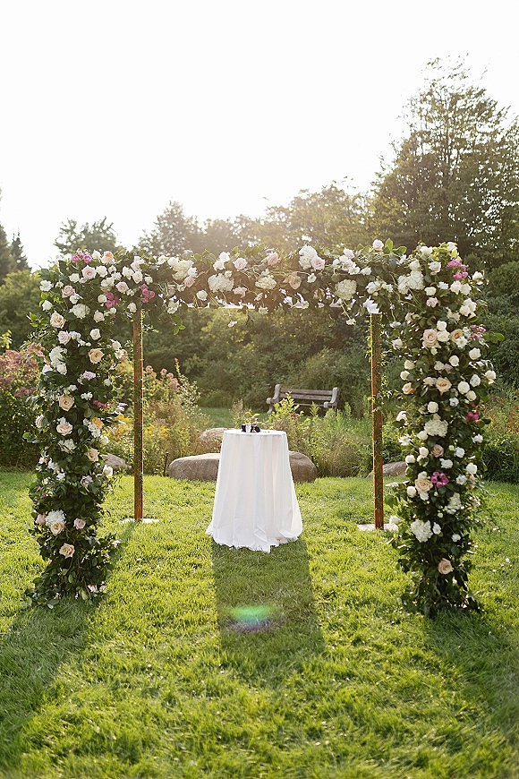 Ceremony arch decor with an outdoor wedding arch of roses, hydrangeas, and greenery on a wooden frame beside a small table on a sunny lawn