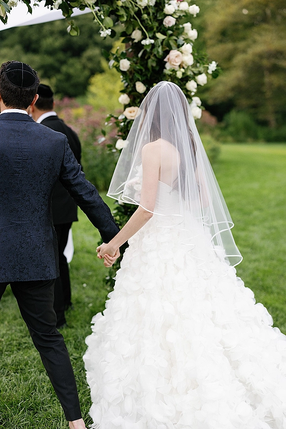 Wedding processional as bride and groom hold hands walking toward a floral arch on an outdoor lawn, her cathedral veil flowing behind
