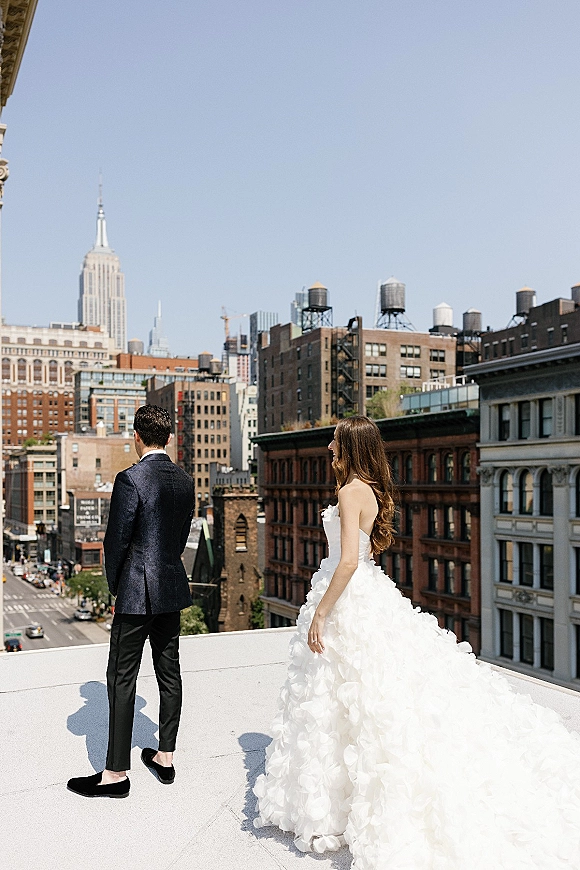 Wedding first look on a rooftop as the bride in a strapless ruffled gown with a long train approaches the groom, city skyline behind