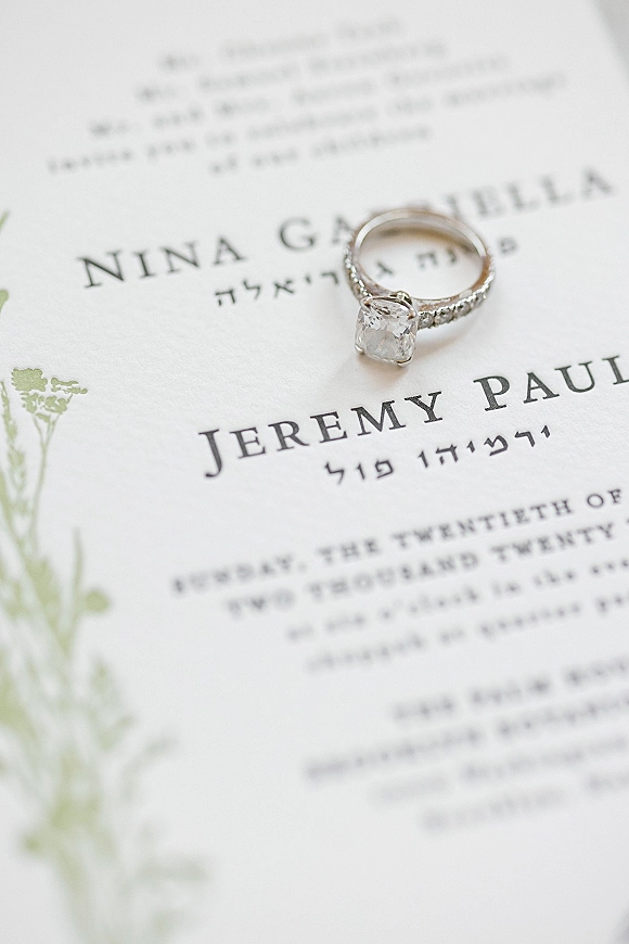 Engagement ring with a pavé band resting on a wedding invitation card, diamond sparkling over botanical illustration and printed text