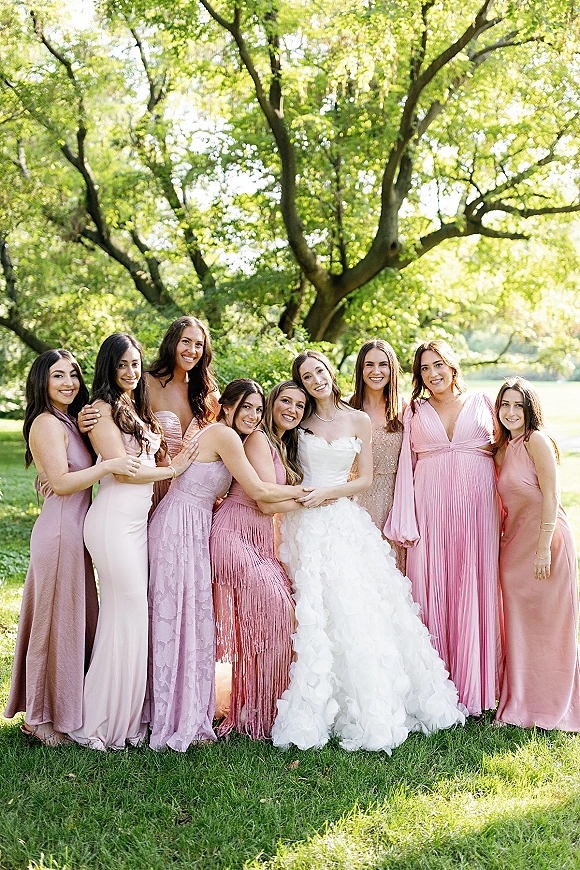 Bridesmaid group photo of the bride with bridesmaids in mismatched pink dresses on a sunny park lawn with large trees and greenery