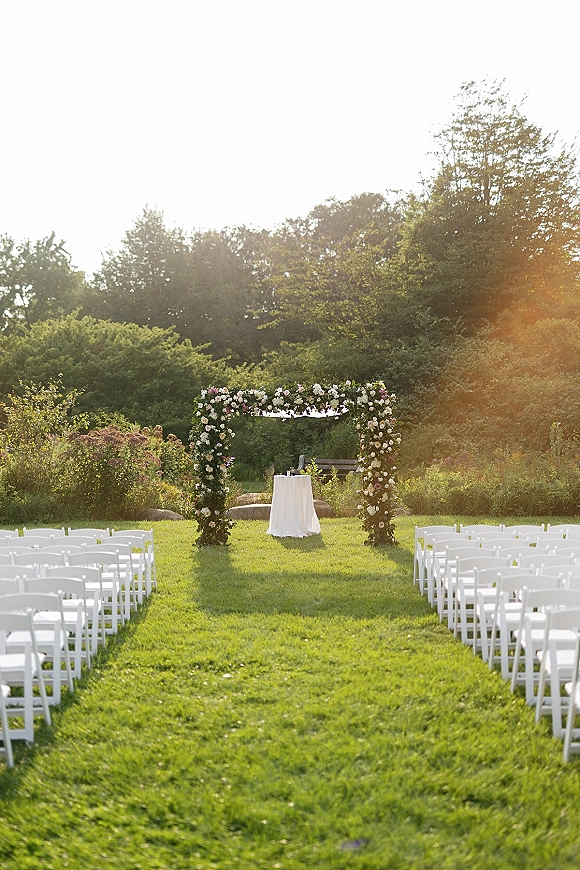 Outdoor ceremony setup with a floral arch and white folding chairs lining the aisle on a sunlit grass lawn with trees and shrubs