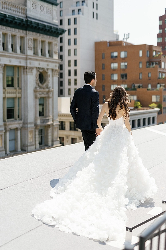 Couple portrait on a rooftop wedding portrait, bride and groom holding hands from behind with a long-train ruffle gown and city skyline behind