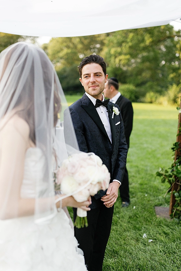 Groom portrait in a black tuxedo, bow tie, and white boutonniere, hands clasped under a greenery wedding arch on a lawn