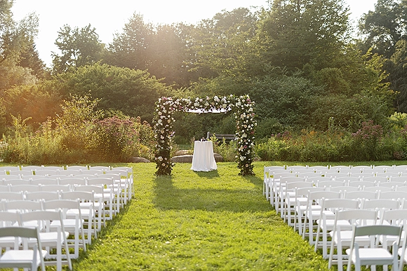 Ceremony setup with floral arch, white aisle chairs and runner leading to an altar table on a sunlit garden lawn with trees