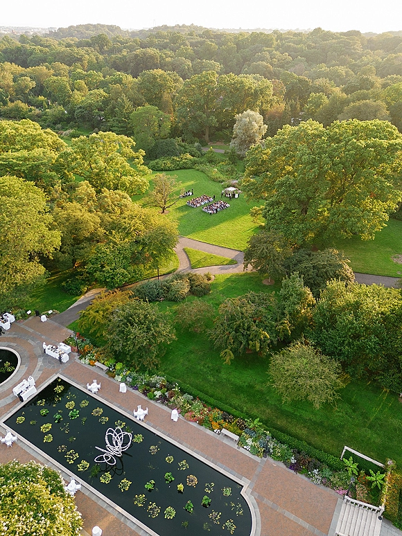 Outdoor wedding ceremony with an aerial wedding ceremony layout, white chairs flanking an aisle to an arch beside a pond and trees at sunset