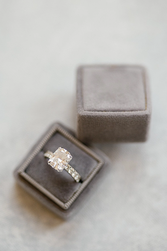 Engagement ring with cushion cut diamond on a pavé band in a gray velvet ring box resting on a clean white surface