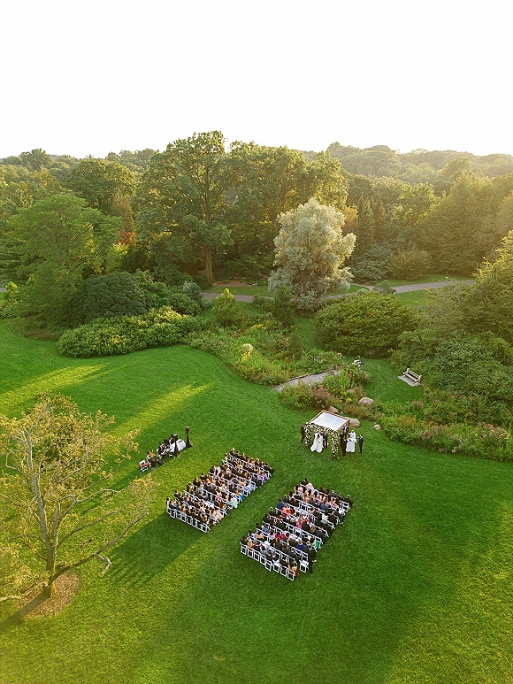 Outdoor wedding ceremony aerial view with white folding chairs facing a floral arch on a sunlit garden lawn with trees and paths