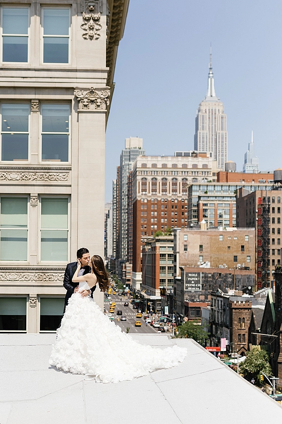 Wedding kiss portrait of bride and groom embracing on a rooftop ledge, her long train flowing, with city skyline and blue sky behind