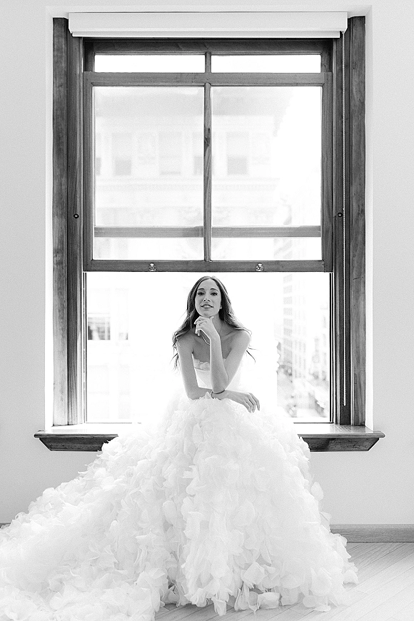 Bridal portrait in black and white of a bride sitting on a windowsill in a strapless gown with a ruffled tulle skirt, city view behind