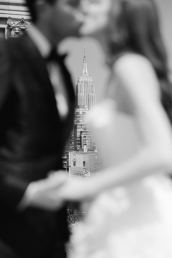 Wedding kiss portrait in black and white, bride in strapless gown and groom in tuxedo kissing with soft-focus city skyline backdrop