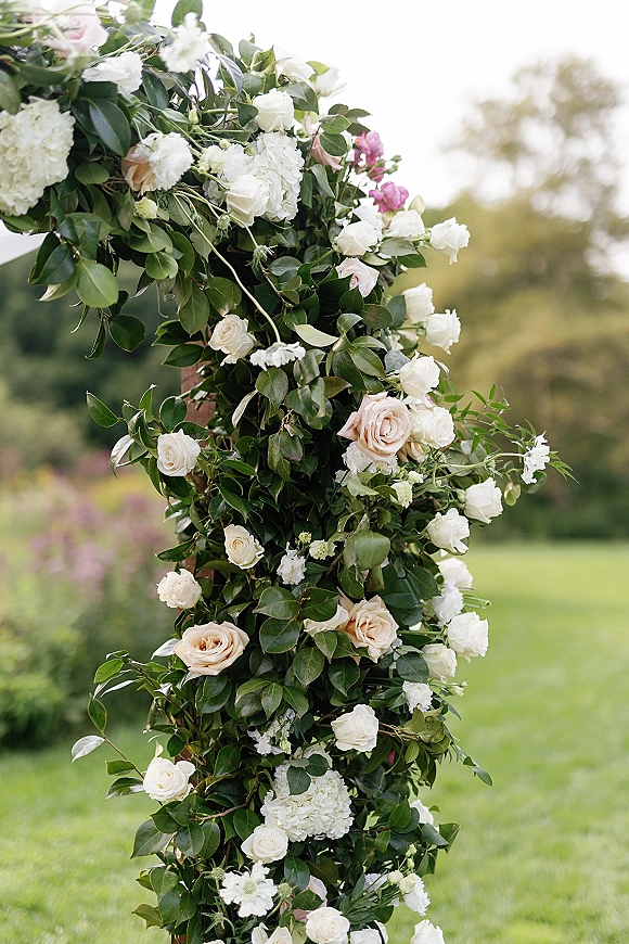 Wedding floral arch with roses and hydrangeas in blush and white, wrapped in lush greenery on a garden lawn with trees and sky