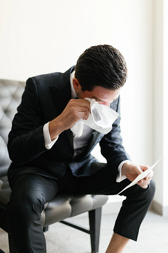 Groom getting ready in a black tuxedo reads a letter card, holding a handkerchief with tie and pocket square in a neutral room