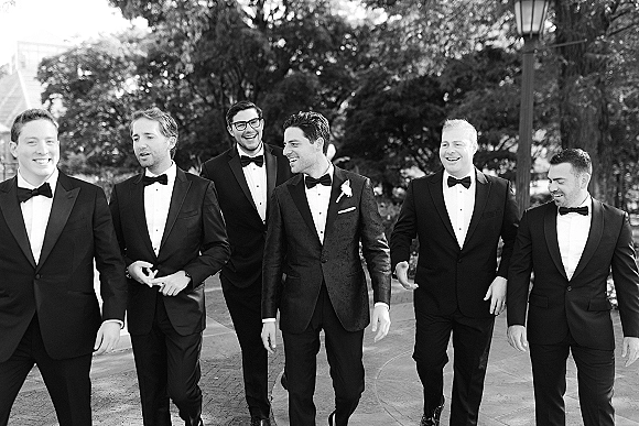 Groomsmen portrait of men walking and laughing in black tuxedos with bow ties on an outdoor walkway lined with trees and a lamppost