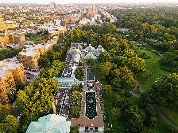 Aerial wedding venue view with a glass greenhouse, reflecting pools and fountains, white chairs on lawns, and city skyline beyond trees