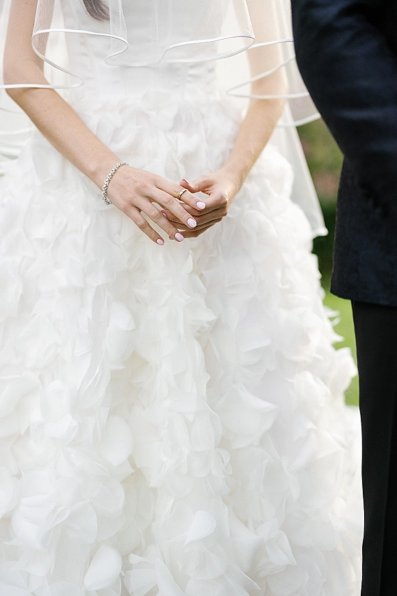 Wedding dress close-up showing ruffle wedding dress texture as bride’s clasped hands display a gold band, bracelet, and pink manicure outdoors