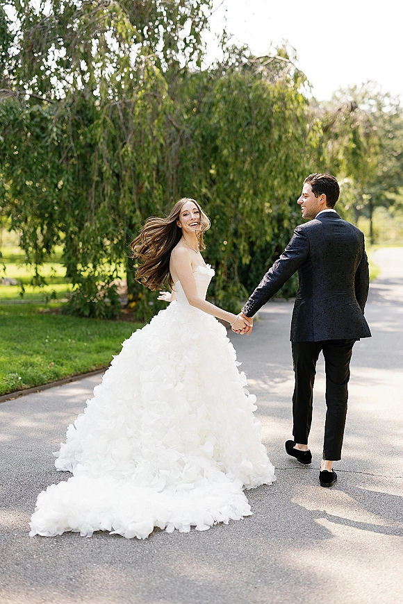 Couple portrait of bride and groom walking hand in hand on a sunlit garden path, her strapless tulle gown train flowing behind