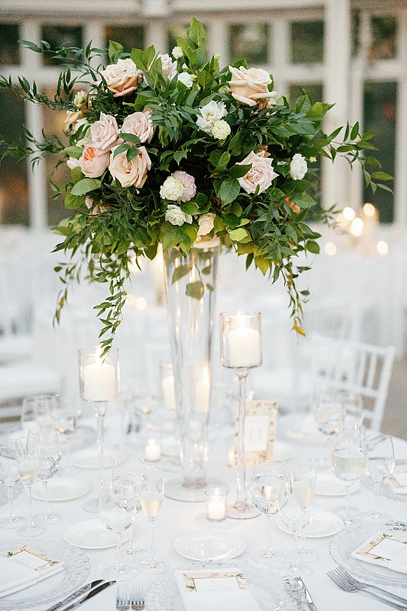 Reception tablescape with wedding tall centerpiece of blush roses and greenery in a clear vase, candlelit place settings under bistro lights