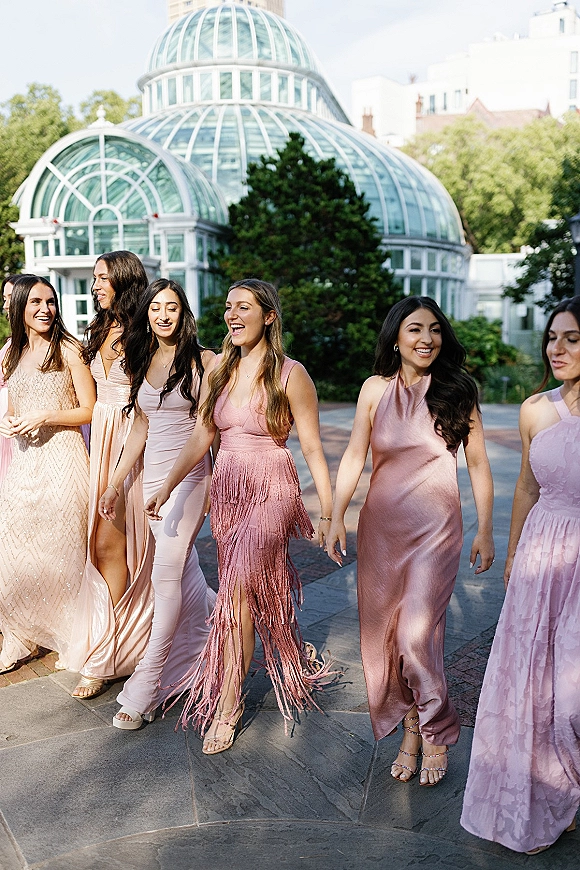 Bridesmaid group photo of friends walking and laughing in mismatched pink bridesmaid dresses along a paved path by a glass conservatory
