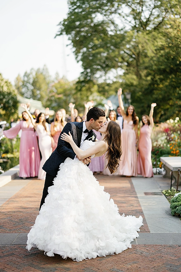 Wedding kiss as the groom dips the bride in a tuxedo and strapless gown, wedding party cheering on a garden stone walkway