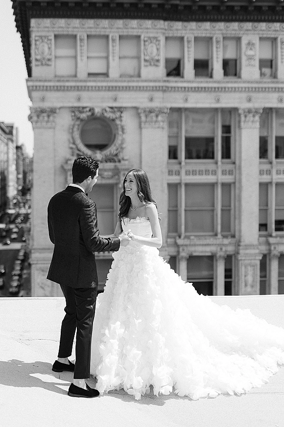Couple portrait in a black and white wedding portrait style, bride in strapless ruffled gown with long train holding hands with groom on rooftop ledge by historic facade