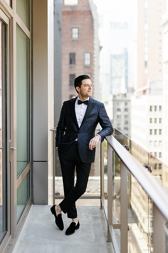 Groom portrait in a black tuxedo and bow tie leaning on a balcony railing, looking away toward city buildings behind him
