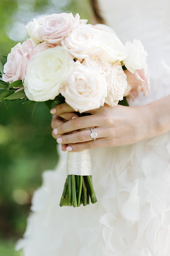 Bridal bouquet of blush roses and white roses with greenery, held in pale pink manicured hands with an engagement ring, bokeh backdrop