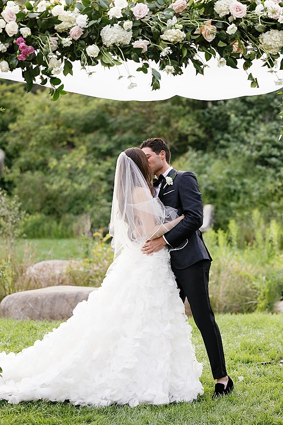 Wedding kiss portrait of bride and groom kissing beneath a rose and hydrangea floral arch, her long veil flowing on a garden lawn