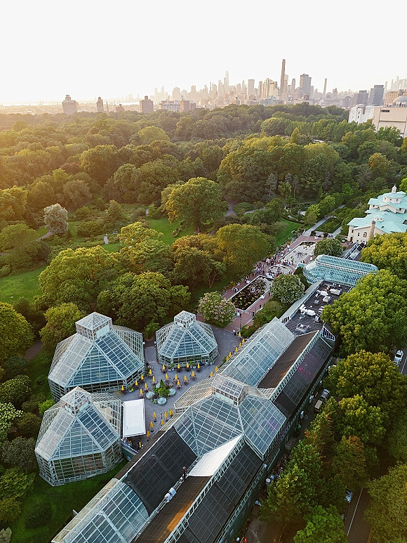 Wedding venue aerial showing a glass greenhouse wedding venue with patio umbrellas, garden paths, tree canopy, and skyline at sunset