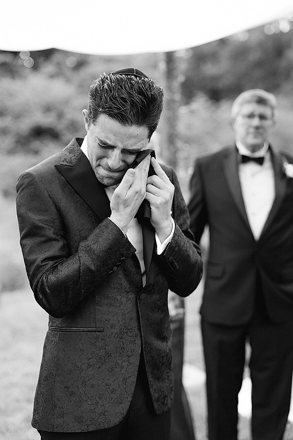 Groom portrait of an emotional groom moment wiping tears in a black tuxedo with boutonniere, trees behind and a blurred guest in background