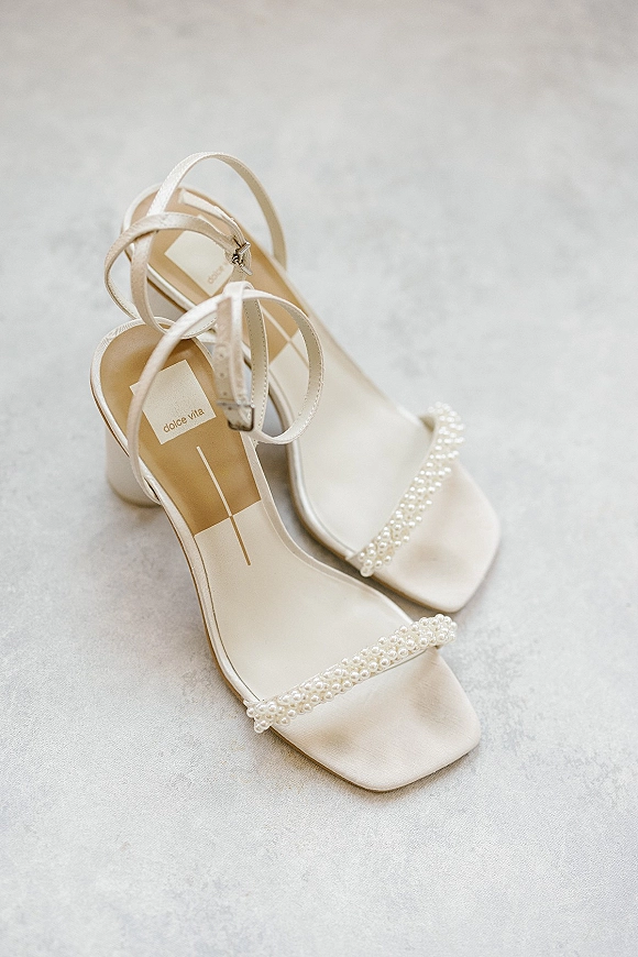 Bridal shoes with pearl wedding heels detailing, ankle straps and block heels on a light gray surface, square toe design