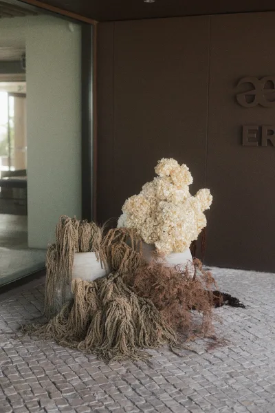 Floral installation of dried wedding flowers with trailing botanicals, arranged on stone pavers by a glass door and brown wall with signage.