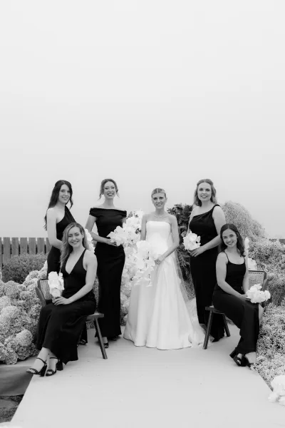 Bride and bridesmaids in a black and white bridal party look, holding white orchid bouquets along a garden aisle by a wooden fence