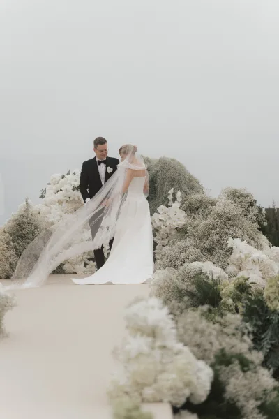 Ceremony moment with bride and groom at altar, her long veil trailing as they face each other under open sky and garden greenery.