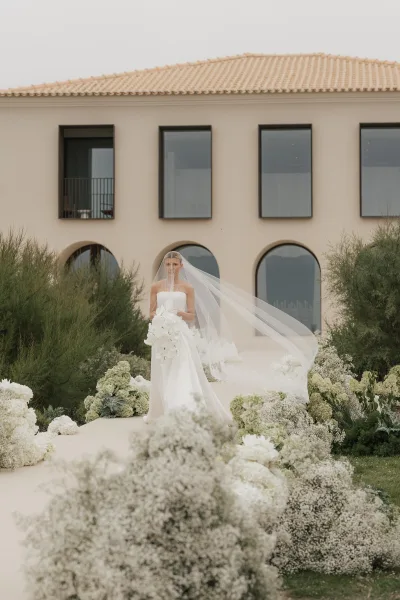 Bridal portrait of a minimalist bride holding a white orchid bouquet before a modern building’s large windows framed by garden shrubs.