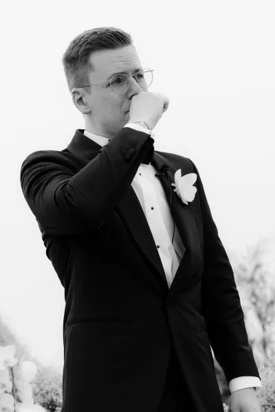 Groom reaction with the groom crying softly behind eyeglasses, framed by a floral backdrop of blooms against open sky and leafy trees.