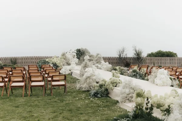 Ceremony aisle design for an outdoor ceremony setup featuring an ivory aisle runner flanked by chairs on a grass lawn before a wooden fence.