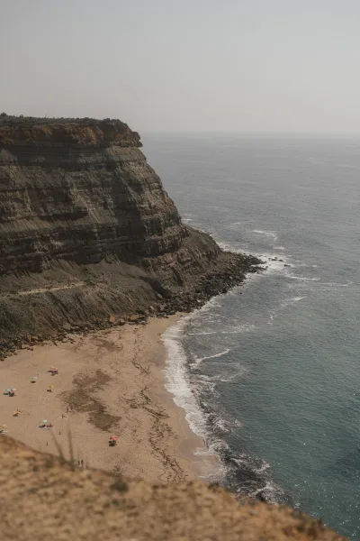 Coastal cliff overlooking a cliffside beach view, with beach umbrellas dotting the shore beneath, set against a tranquil ocean horizon.