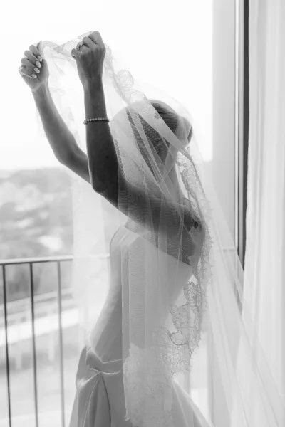 Bridal portrait of a bride getting ready by a window, a lace veil catching natural light as curtains frame a balcony railing and landscape.