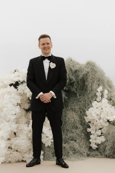 Groom portrait showcasing a tuxedo groom look with a classic bow tie, framed by a white floral installation and lush greenery under open sky.