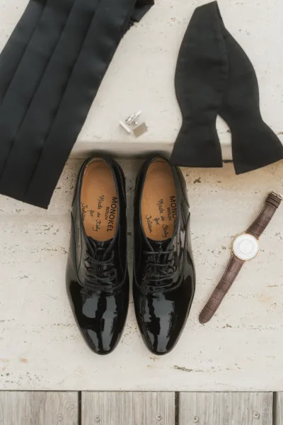 Groom accessories with black patent oxford shoes and an untied black bow tie arranged on a stone surface with wood planks