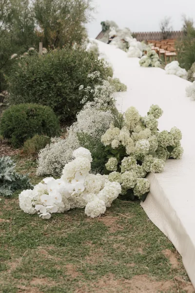 Ceremony aisle flowers in lush hydrangea aisle decor, creating low meadow-like arrangements on a grass lawn with garden shrubs and wooden chairs.