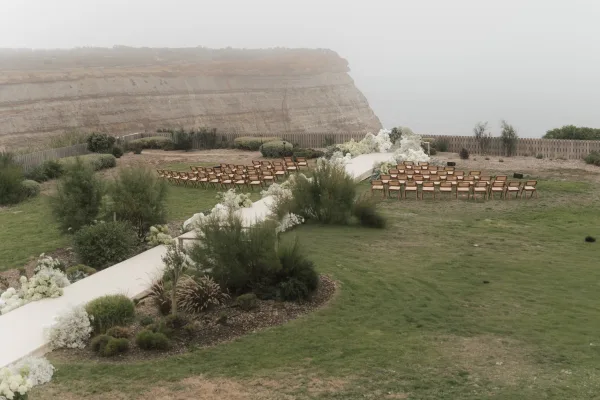 Ceremony setup on a cliffside lawn with outdoor ceremony decor, white florals lining the aisle as the ocean fades into a foggy sky.