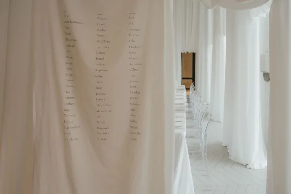 Wedding seating chart styled as a fabric seating chart banner, framed by white curtains over a light herringbone floor near a doorway