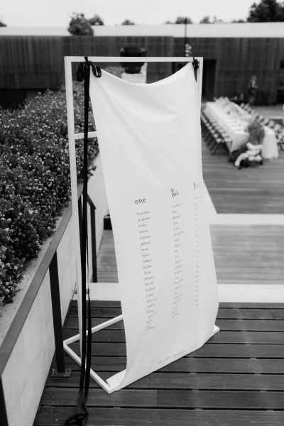 Wedding seating chart on a fabric seating chart, suspended from a metal frame on a wood deck beside banquet tables and a trimmed garden hedge.