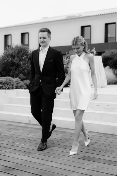 Couple portrait of a bride and groom walking hand in hand, the bride in a bridal mini dress, on stone steps by a wooden deck and shrubs.