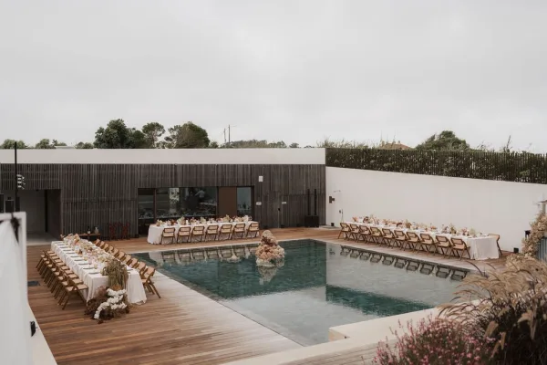 Outdoor reception setup for a poolside wedding reception, featuring long banquet tables with white linens and dried florals.