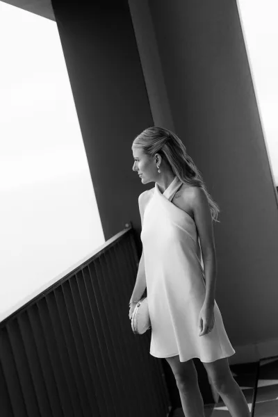 Bridal portrait of a little white dress bride in a one-shoulder mini dress by a balcony railing, softly lit by window light.