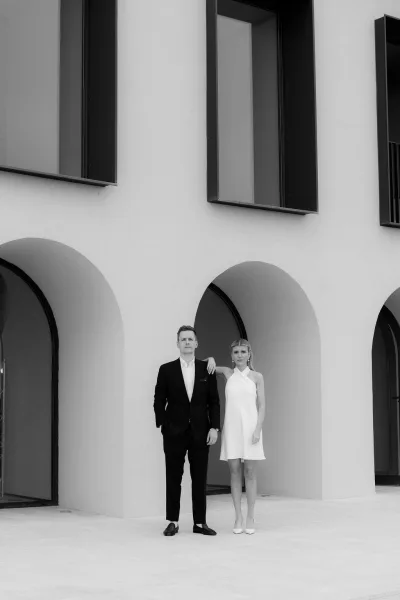Couple portrait, a black and white wedding portrait with the bride in a bridal mini dress before an arched stucco facade with large windows.